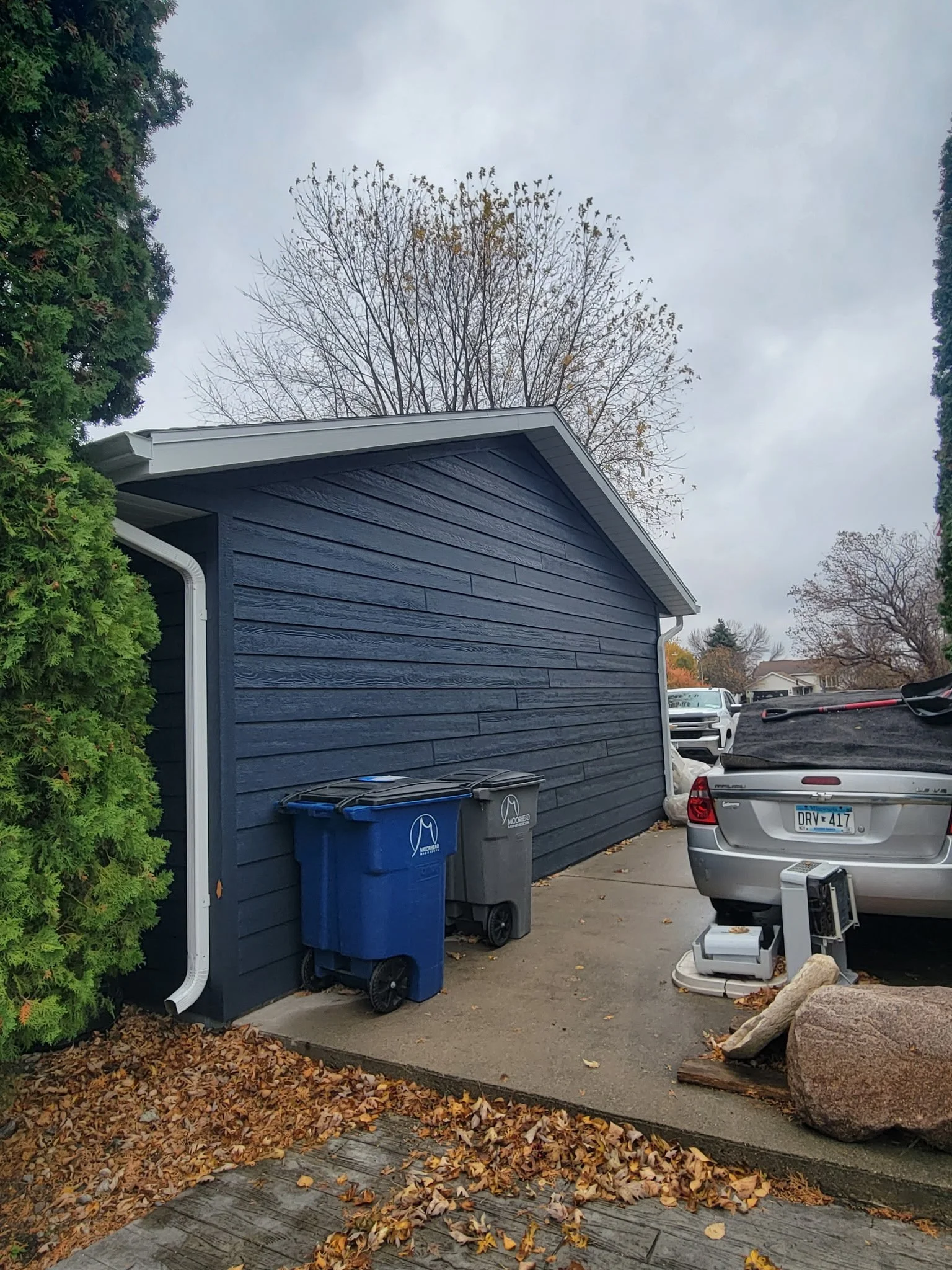 View of a house with dark blue siding, two trash bins (blue and gray) beside it, a car parked on the driveway with a computer and a small printer in front, fall leaves on the ground, and trees with bare branches in the background on a cloudy day.