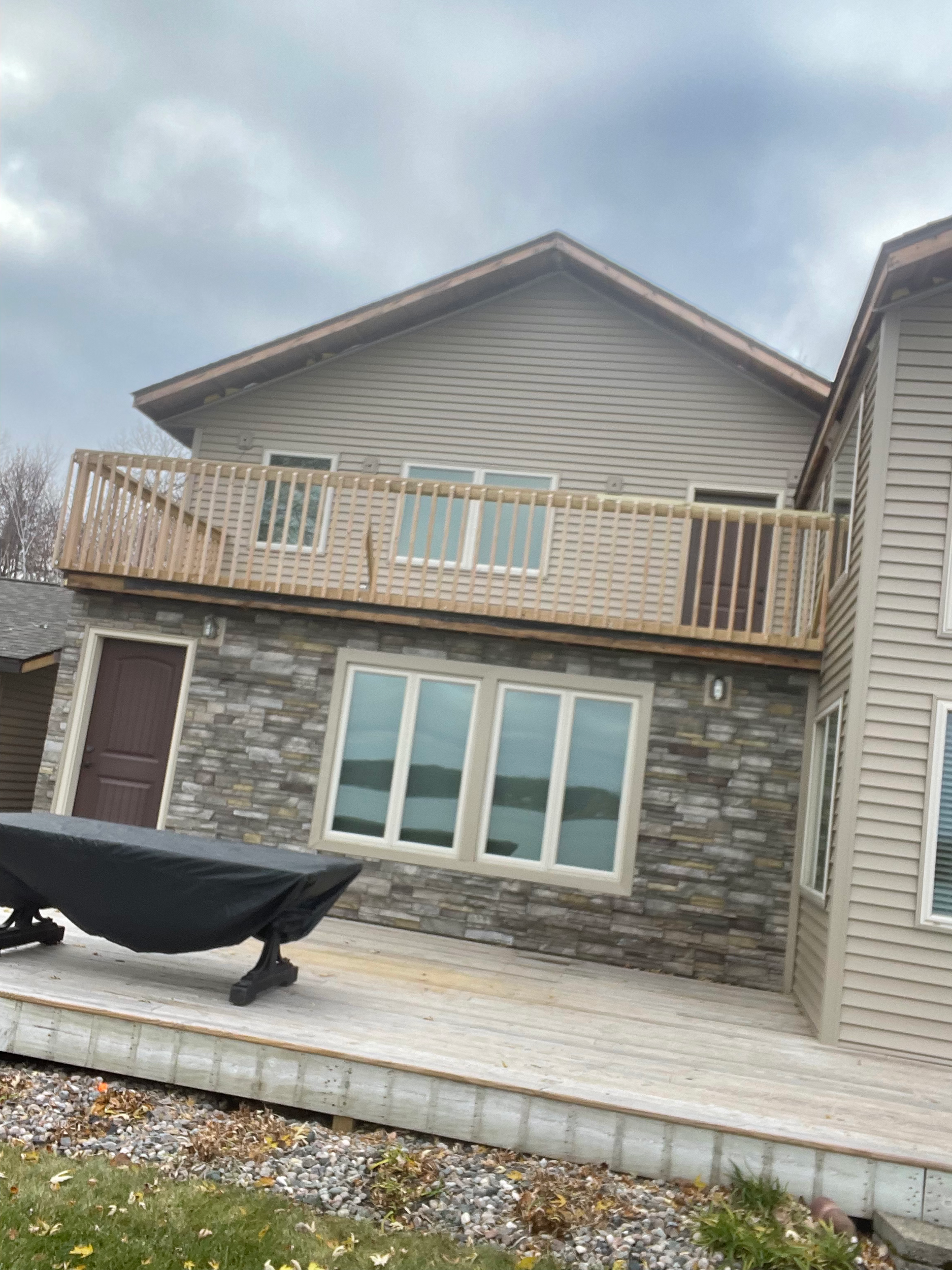 Backyard with a wooden deck, sliding glass doors, and a small balcony with a railing. There is a grill covered with a black cover on the deck and a garden bed with rocks on the right side. The house has beige siding and stone accents.