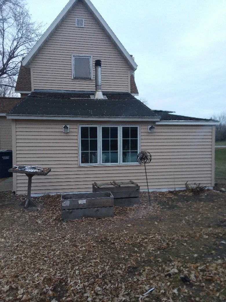 Backyard of a house with beige siding, a large window, and a small attic window. There is a chimney pipe on the roof, and some gardening tools and wooden planters on the ground.
