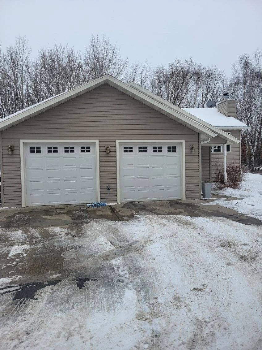 A beige, two-car garage with white doors, surrounded by snow and ice in a winter setting, with leafless trees in the background.
