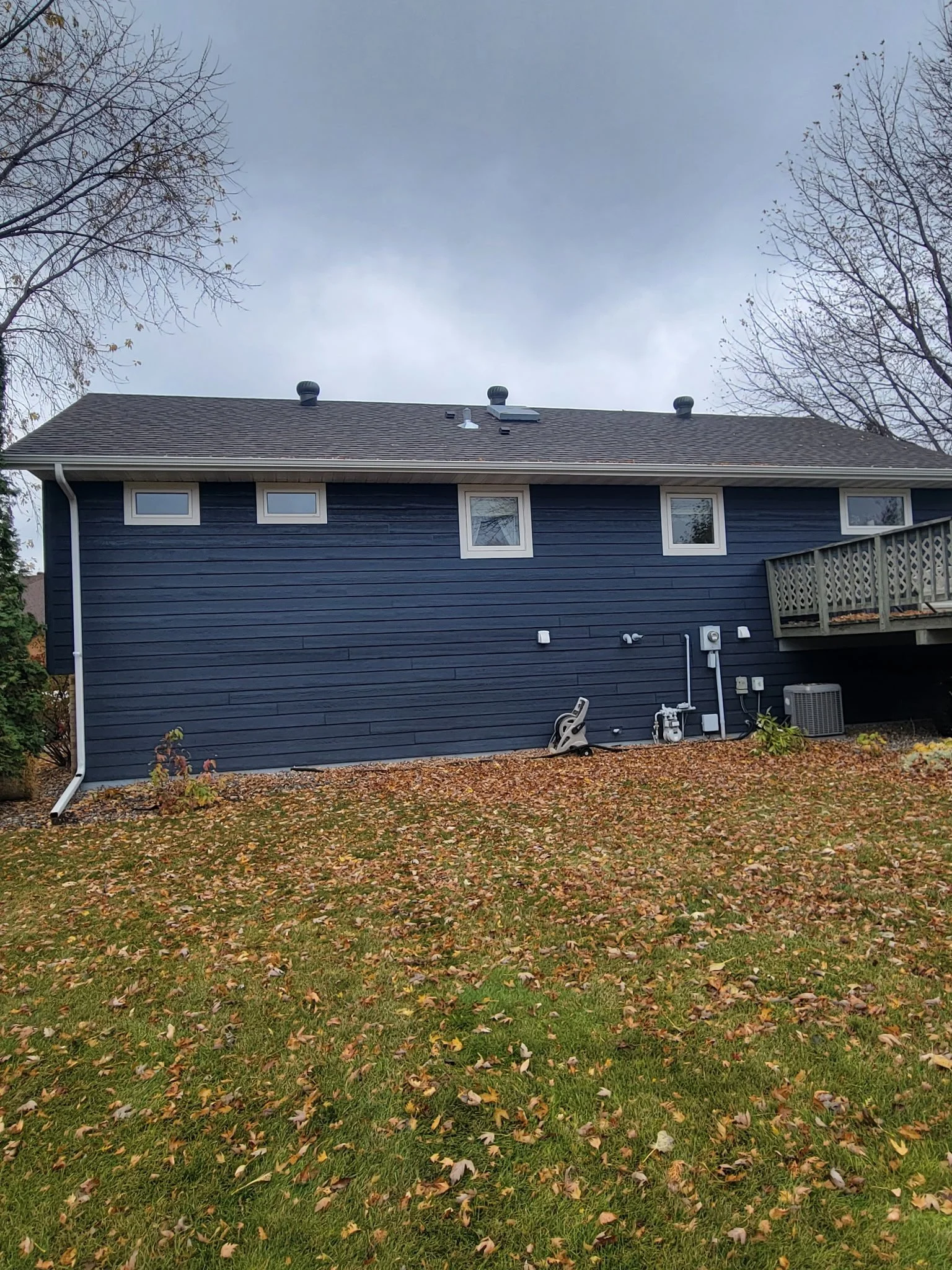 Backside of a blue house with a deck, several small windows, and outdoor equipment, surrounded by fallen leaves and a grassy yard.