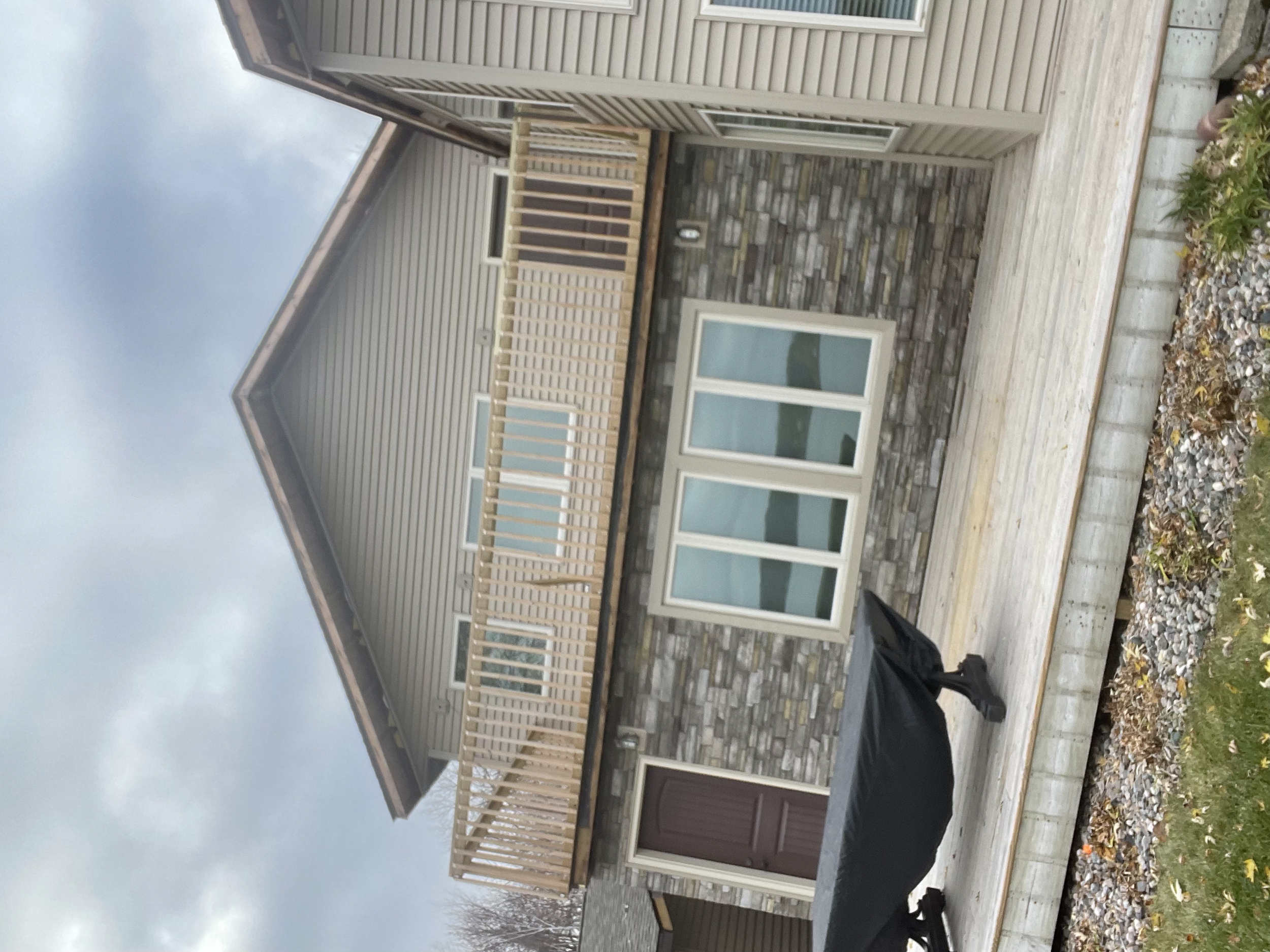 Backyard with a wooden deck, sliding glass doors, and a small balcony with a railing. There is a grill covered with a black cover on the deck and a garden bed with rocks on the right side. The house has beige siding and stone accents.