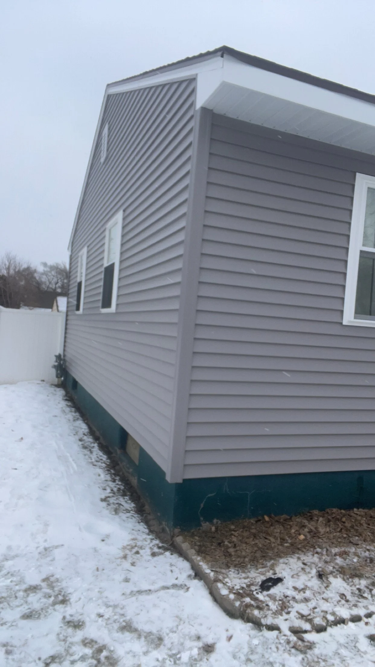 Side view of a house with gray vinyl siding, white trim, and snow on the ground.