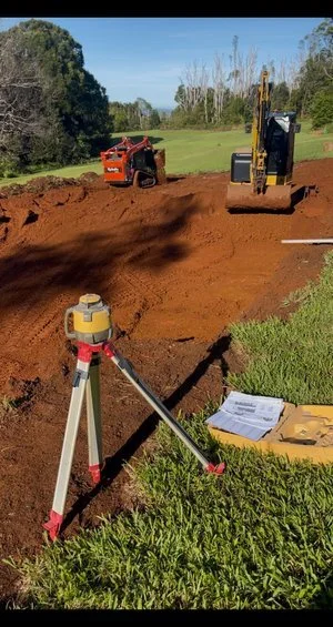 Construction site with leveling equipment, a compact track loader, and a small excavator on red soil, surrounded by grass and trees. Blueprints are visible in the foreground.