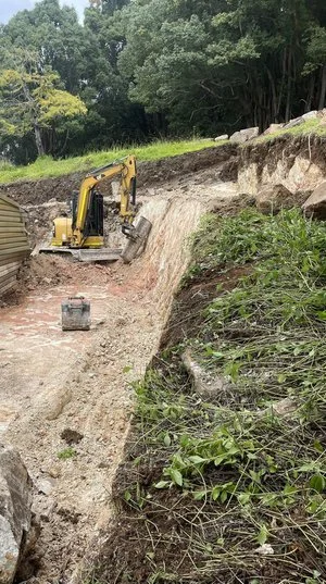 Excavator digging a trench near a forested area with a slope covered in grass and rocks.