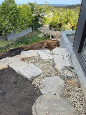 Stone pathway under construction next to a building, with dirt and gravel, surrounded by trees.