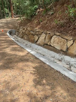 A stone retaining wall along a dirt path in a wooded area with a concrete drainage ditch at the base.