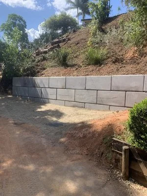 Concrete retaining wall on a hillside with plants and trees around, under a clear blue sky.