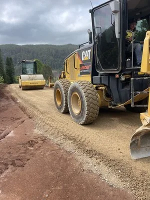Construction site with heavy machinery, including a road grader and a steamroller, working on a gravel road amidst a hilly, forested area under a cloudy sky.