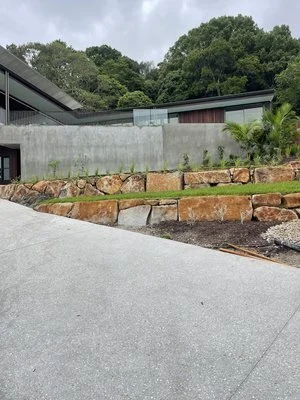 Modern house with concrete walls and a stone retaining wall, surrounded by greenery.
