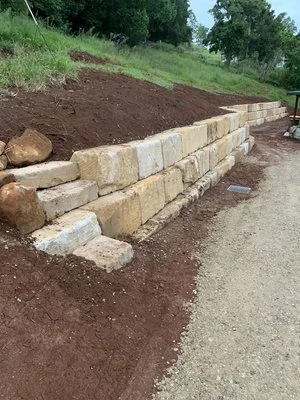 Stone retaining wall on sloped terrain with fresh soil and grassy area in the background.