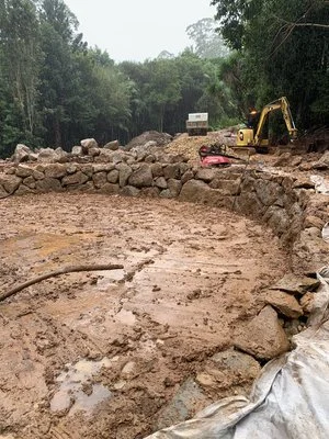 Construction site with a muddy area, rocks, and an excavator near a forested area.