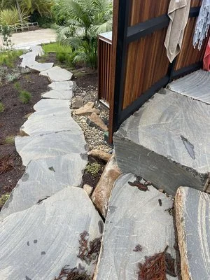 Outdoor stone pathway with large flat stones, surrounded by plants and a wooden fence on the right.