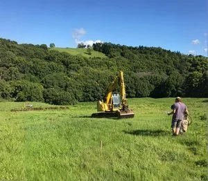 Site survey with man walking towards a yellow excavator in a grassy field with forested hills in the background.