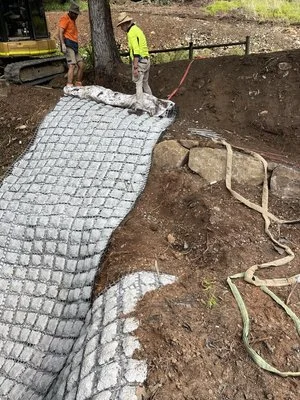 Workers installing a flexible concrete mat on a slope for erosion control, with a construction vehicle nearby.