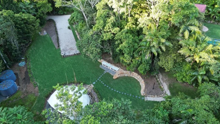 Aerial view of a landscaped garden with a grassy area, stone pathways, and dense surrounding trees. Features include a white dome structure, tanks, and a wooden bench.