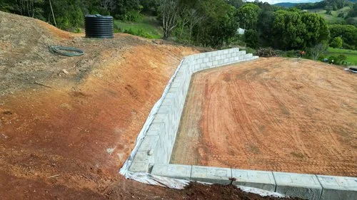 Retaining wall under construction on a sloped terrain with soil and grass, surrounded by rural landscape.