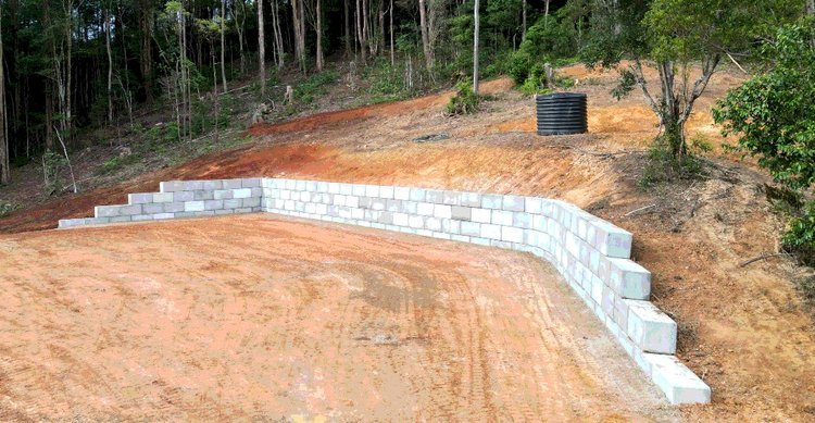 Retaining wall made of concrete blocks on a sloped dirt area with surrounding trees.