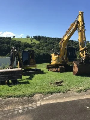 Two yellow excavators on grassy field with trees and hills in the background under a clear blue sky.