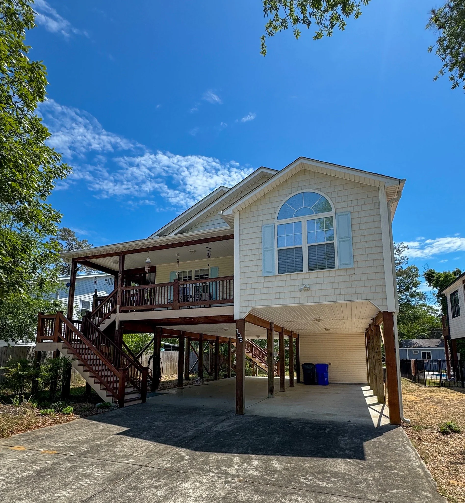 A two-story house with light-colored siding, an arched window, and a wooden deck with stairs on the left side. The house has a covered parking area underneath and is surrounded by trees and a clear blue sky with some clouds.