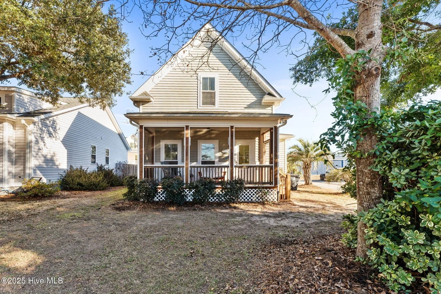 A two-story house with a screened-in front porch, light gray siding, white trim, and a gable roof. The yard has some bushes, a large tree on the right, and patches of grass and dirt.