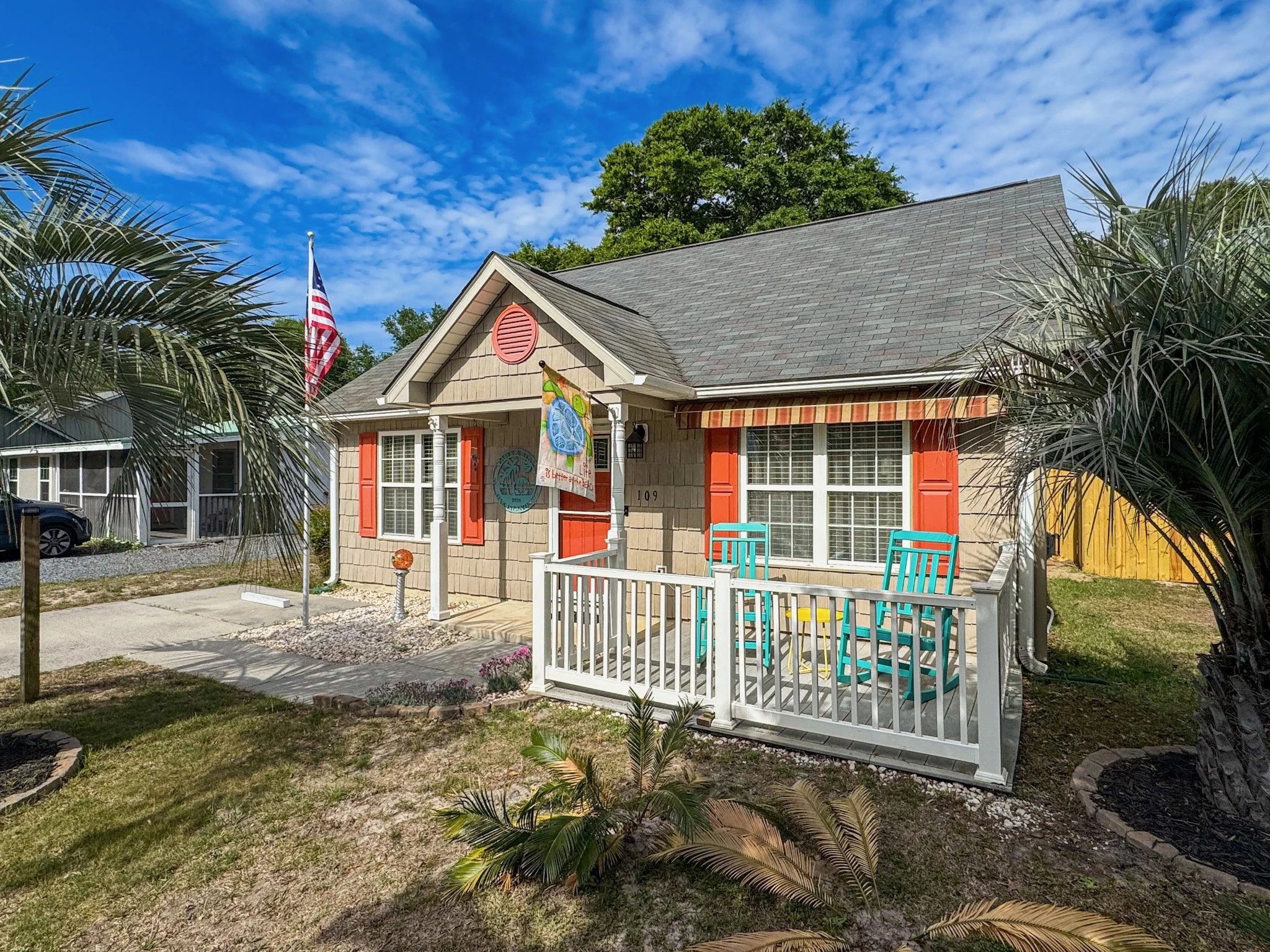 Front view of a house with a gray roof, beige walls, orange shutters, white porch, and a colorful flag, surrounded by palm trees and greenery, under a blue sky with scattered clouds.