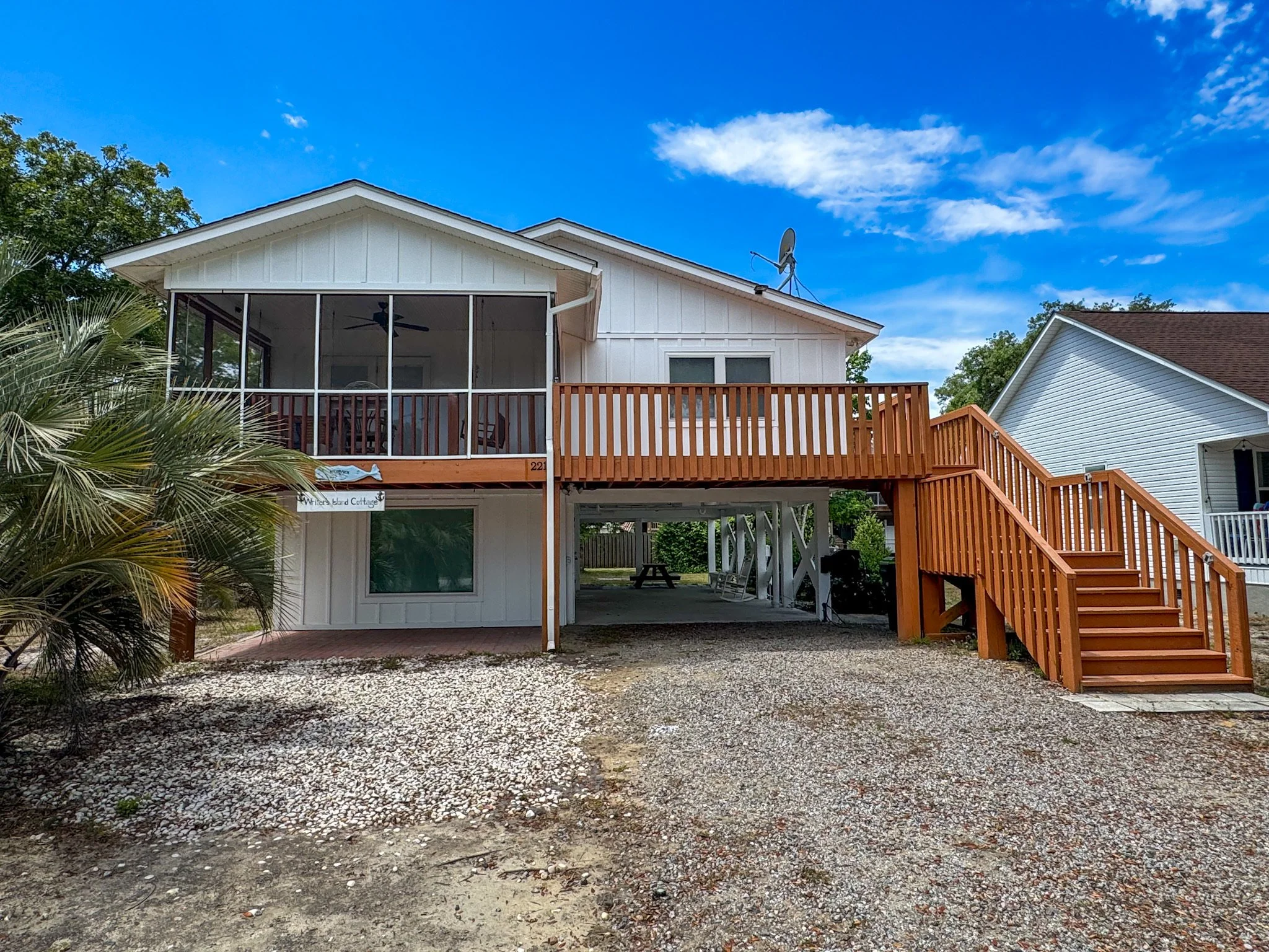 View of the back of a two-story white house with a wooden deck, staircase, and a screened porch, under a blue sky with some clouds, and a gravel yard in the foreground.
