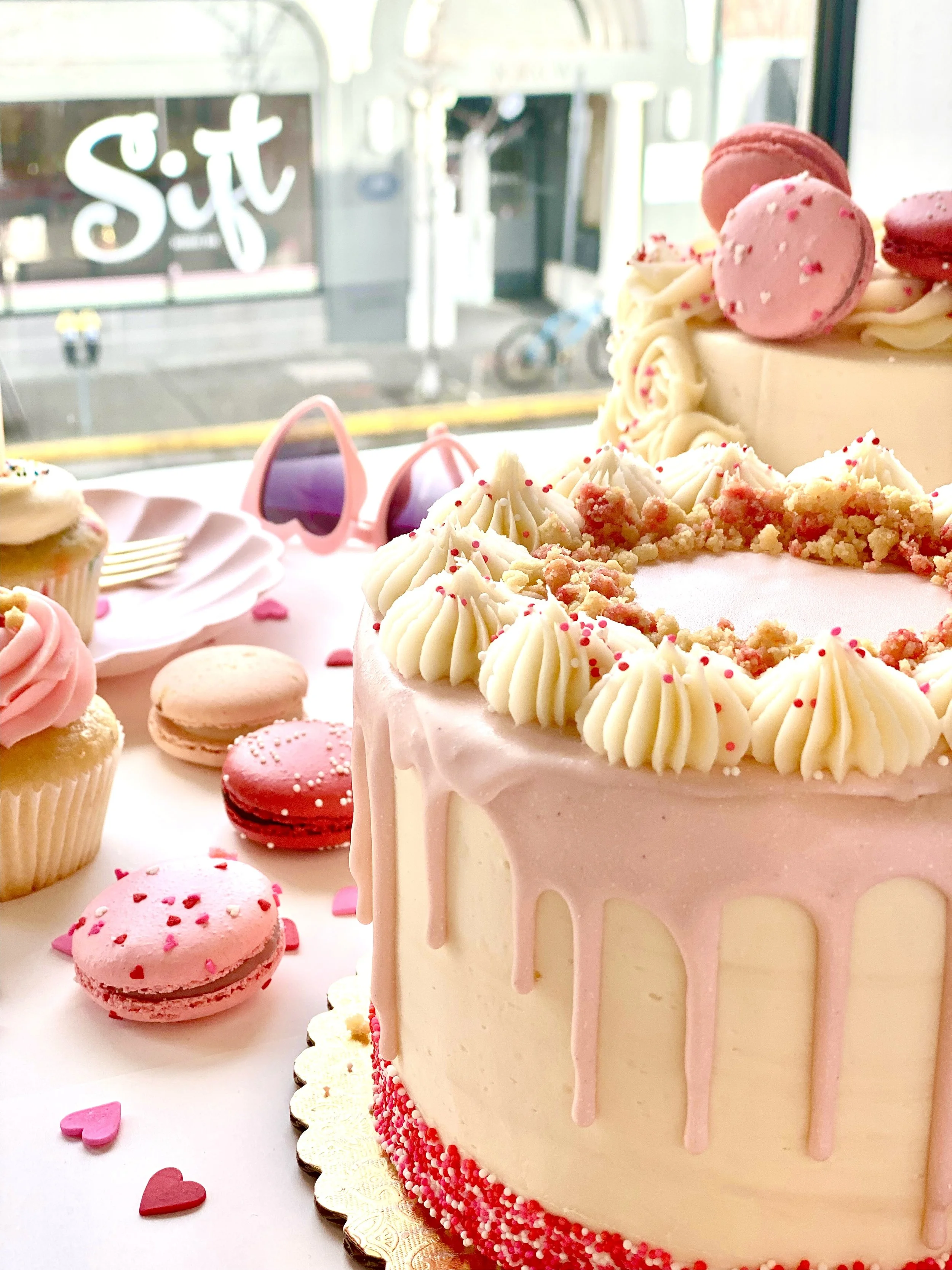A pink and white birthday cake with pink macarons and white icing decorations, surrounded by pink macarons, sprinkles, and small heart-shaped candies on a table with a window view outside.