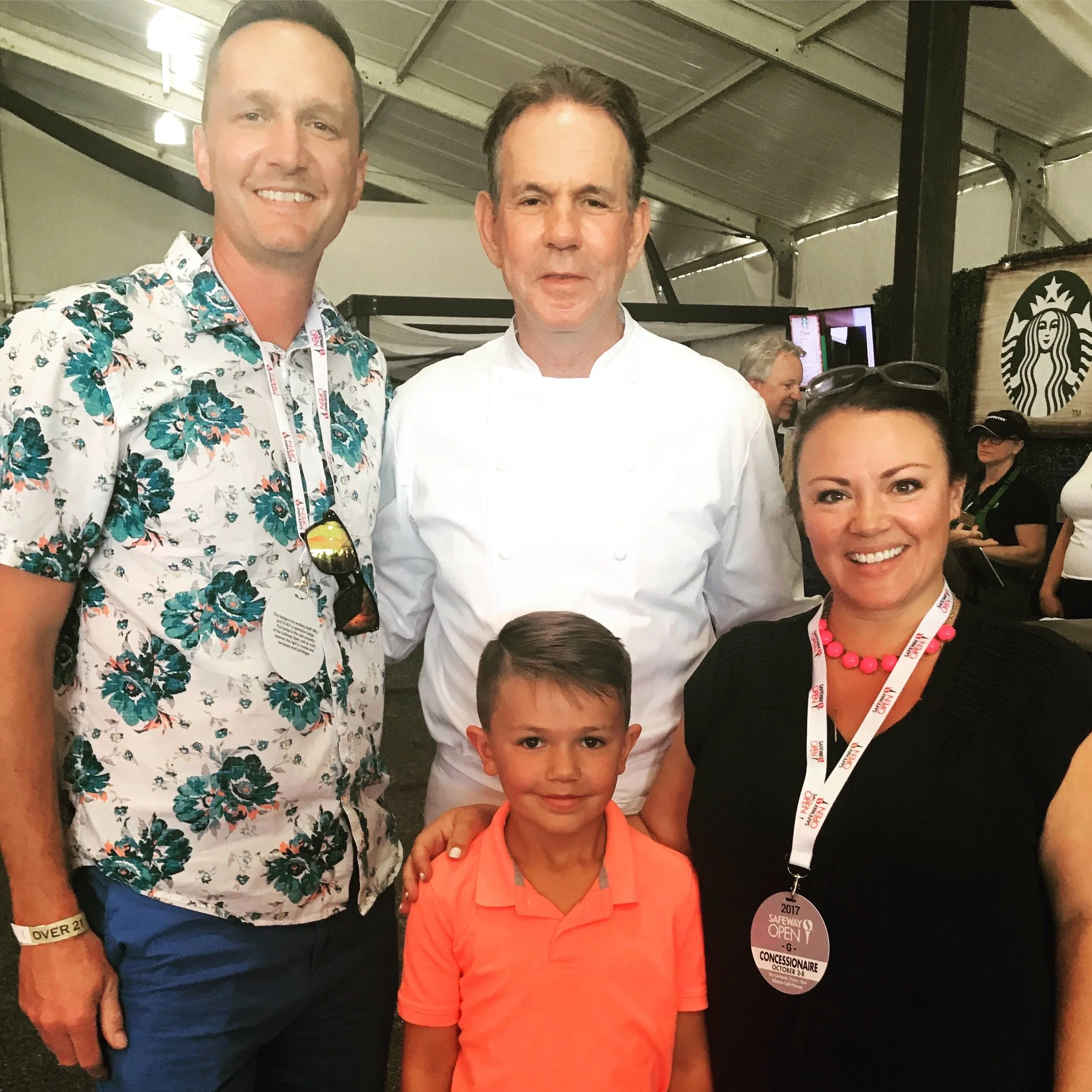 Group of four people and a young boy at an event, with a man in a white chef's coat in the center. The others are a man in a floral shirt, a woman in black with pink necklace and sunglasses on her head, and the boy in an orange shirt. They are inside