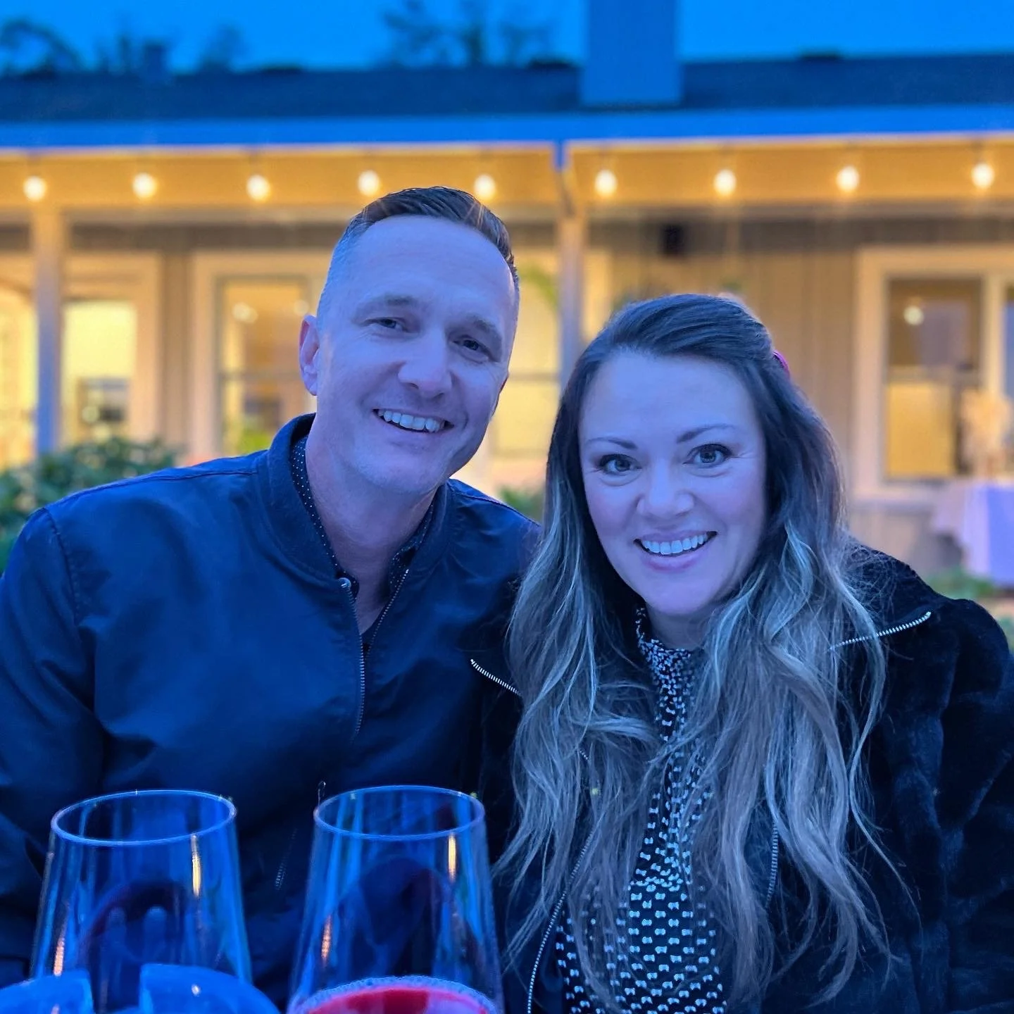 The Balluses. sitting together outdoors at night, with glasses of wine in front of them, in front of a house with string lights.