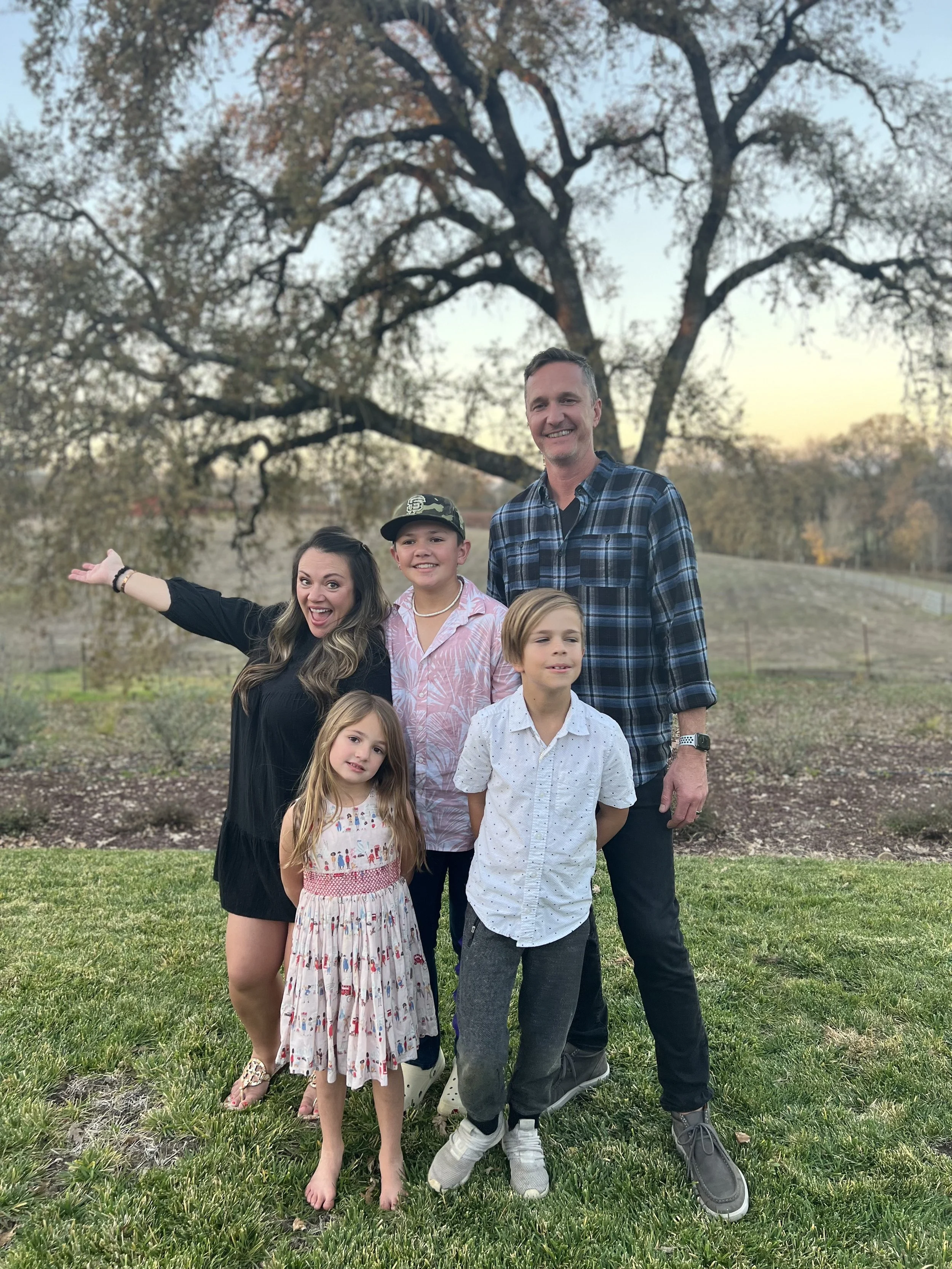Family of six outdoors during sunset, standing on grass with a large tree in the background.