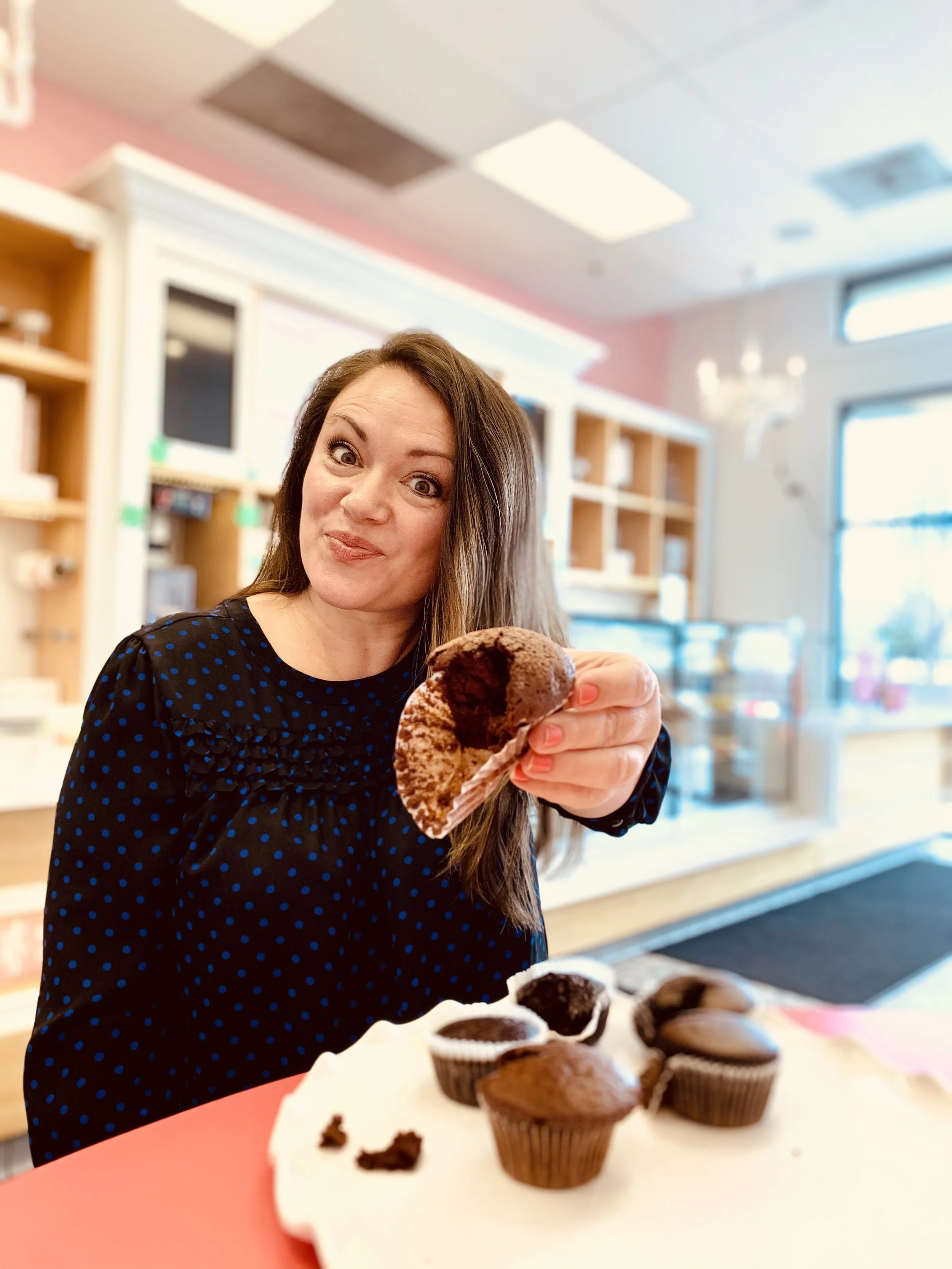 Woman with long brown hair in a black dress with blue polka dots holding a chocolate cupcake inside a bakery, with a plate of various cupcakes in front of her.