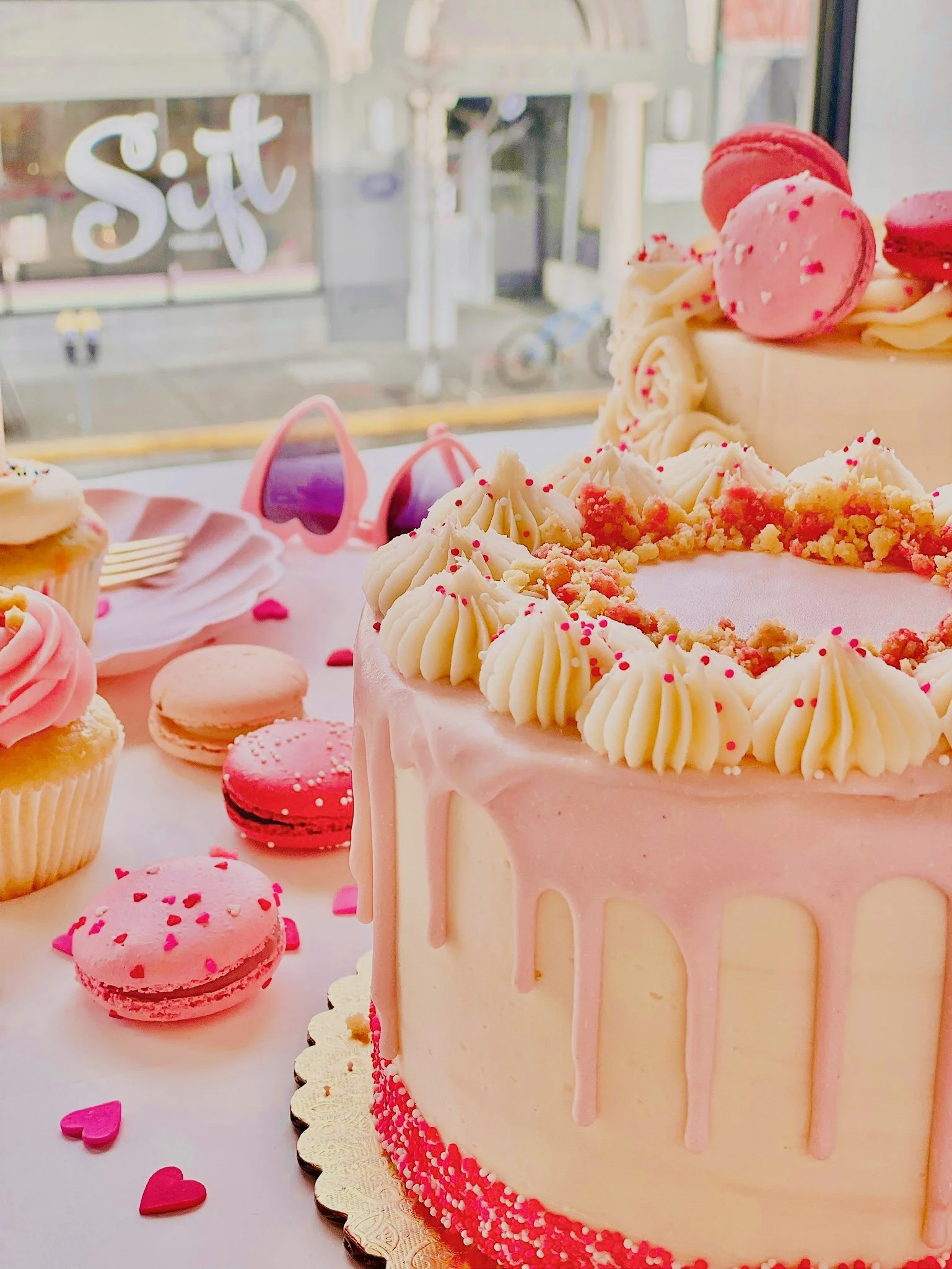 Valentines at Sift Dessert Bar. A pink and white decorated birthday cake with pink macarons on top, surrounded by pink cupcakes, macarons, and small heart-shaped decorations on a table with a window in the background.