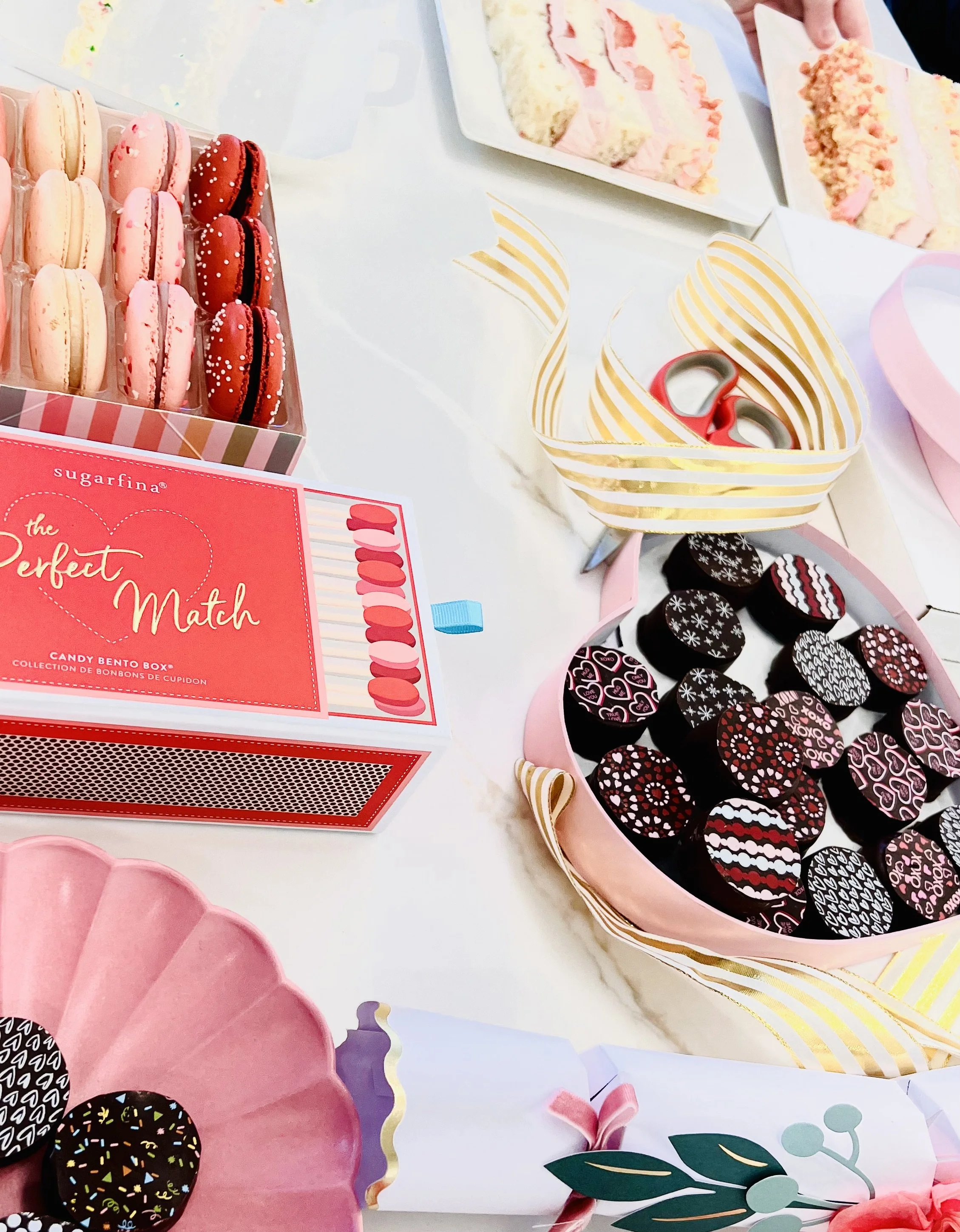 Assorted Valentine-themed candies including pink, red, black and white chocolates with decorative patterns, macarons, and candy boxes on a white table. By Sift Dessert Bar