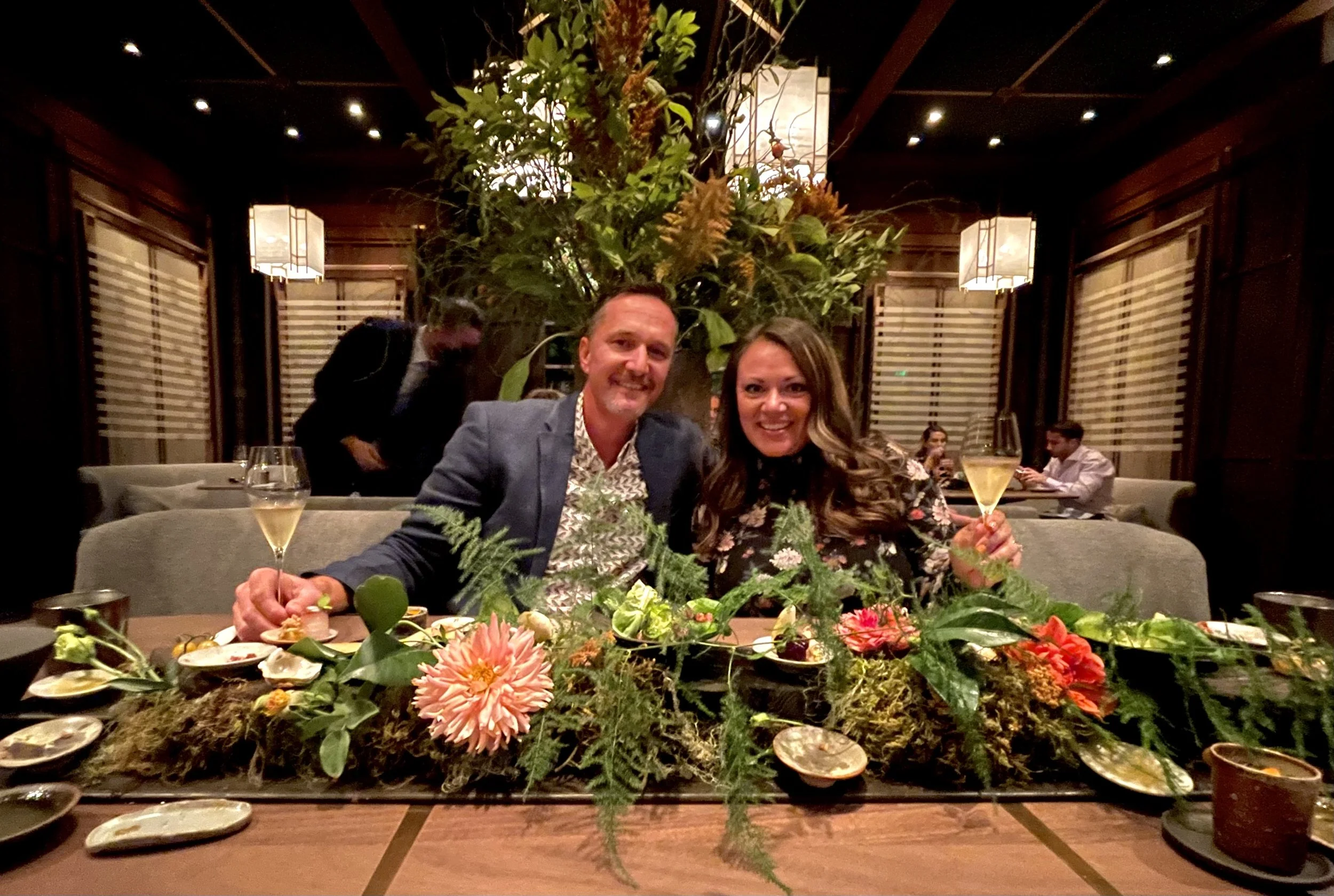 A man and woman sitting at a dining table with a floral centerpiece, both smiling and holding champagne glasses, in a restaurant setting.