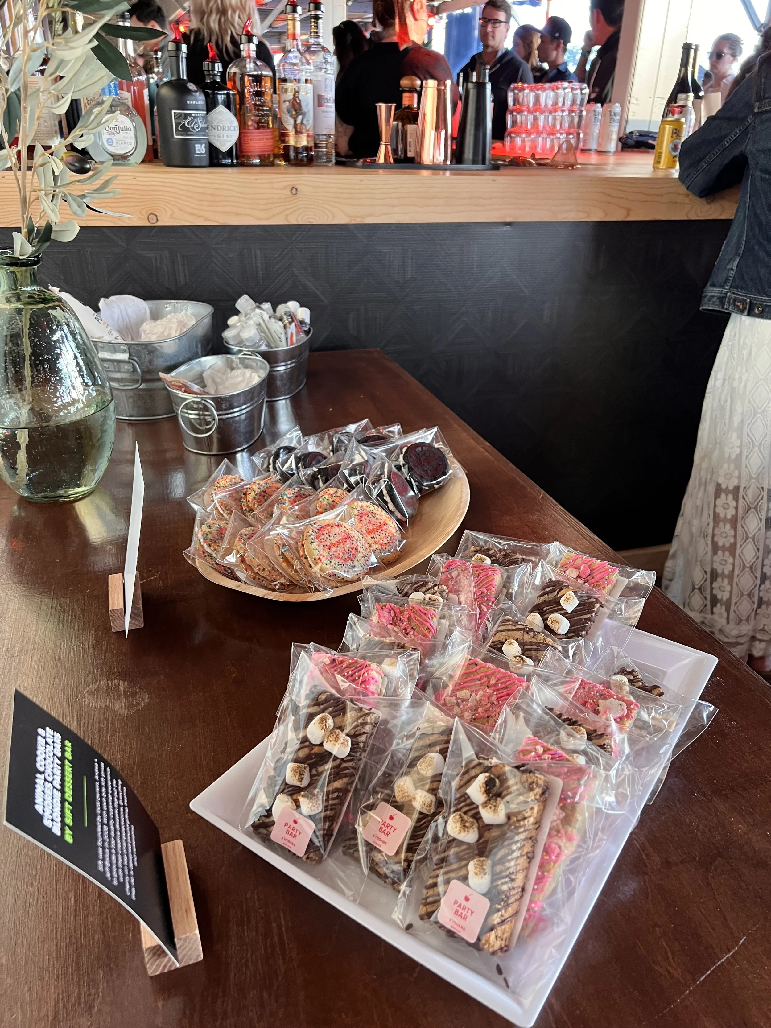 Assorted decorated cookies and treats on a table at a party or event, with a bar area and people in the background.