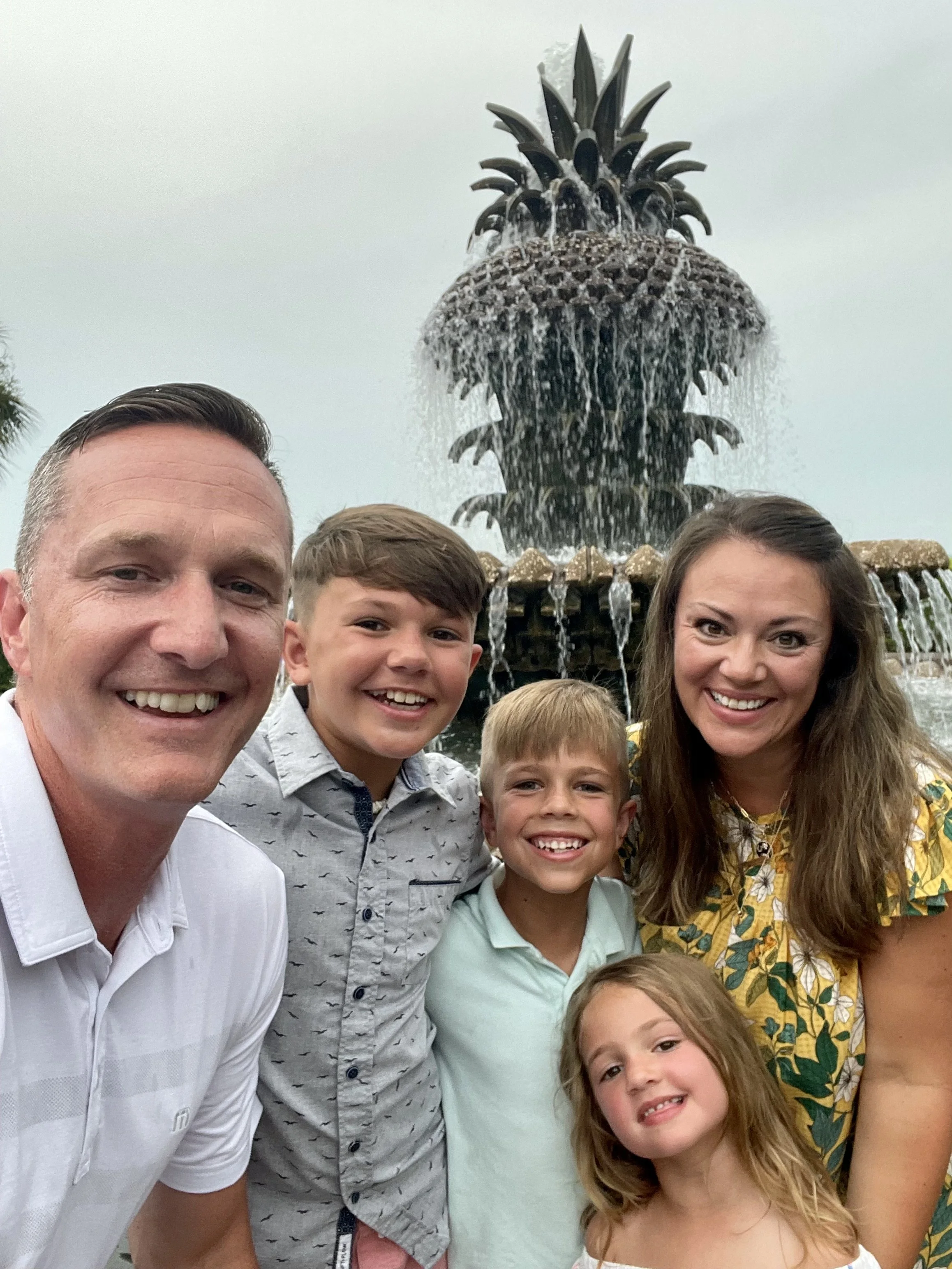 A family of five taking a selfie in front of a pineapple-shaped fountain with water flowing from its top, outdoor setting during daytime.