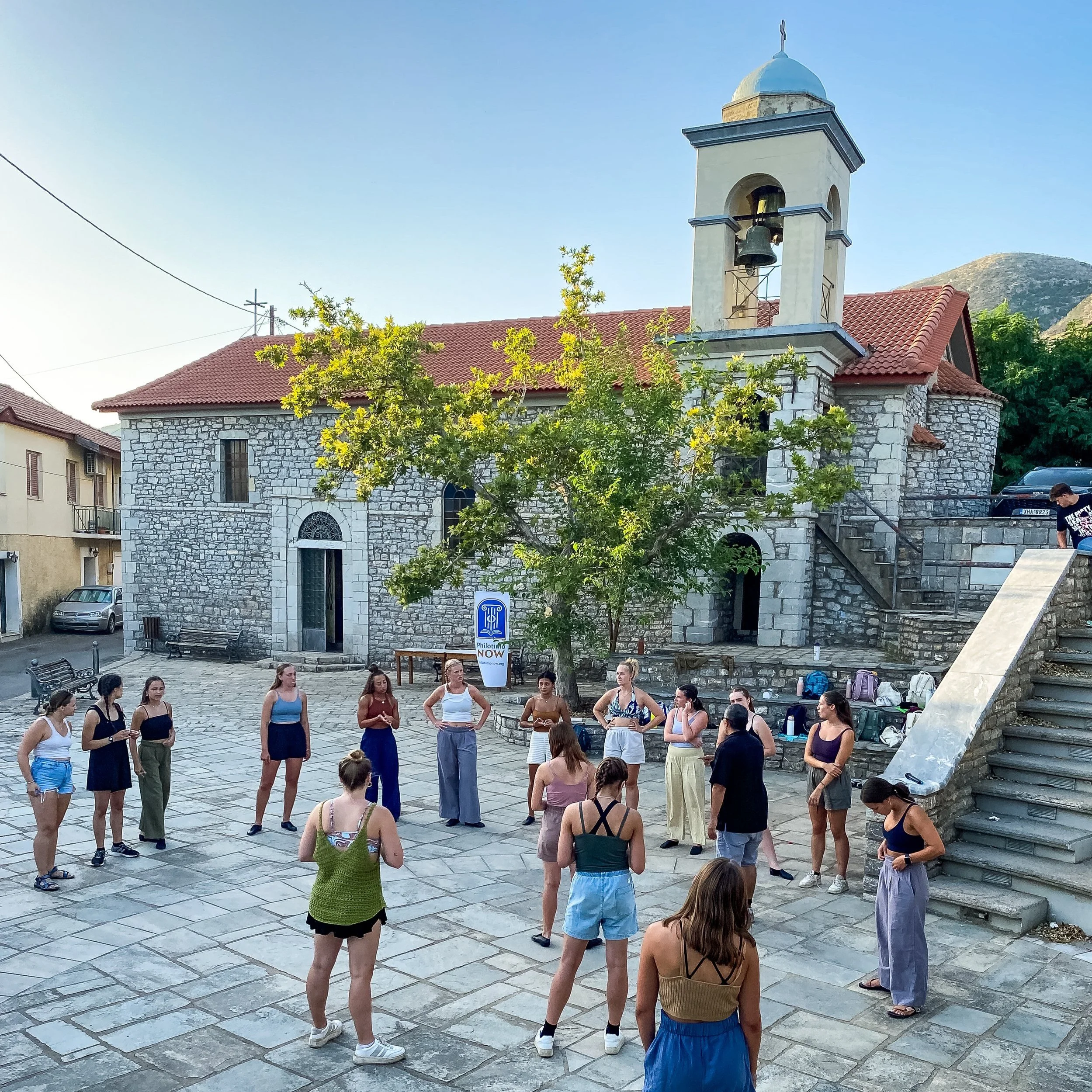 a group of students stand in a courtyard square in front of a stone church with a bell tower as part of a literacy scholar trip to greece from shenandoah university