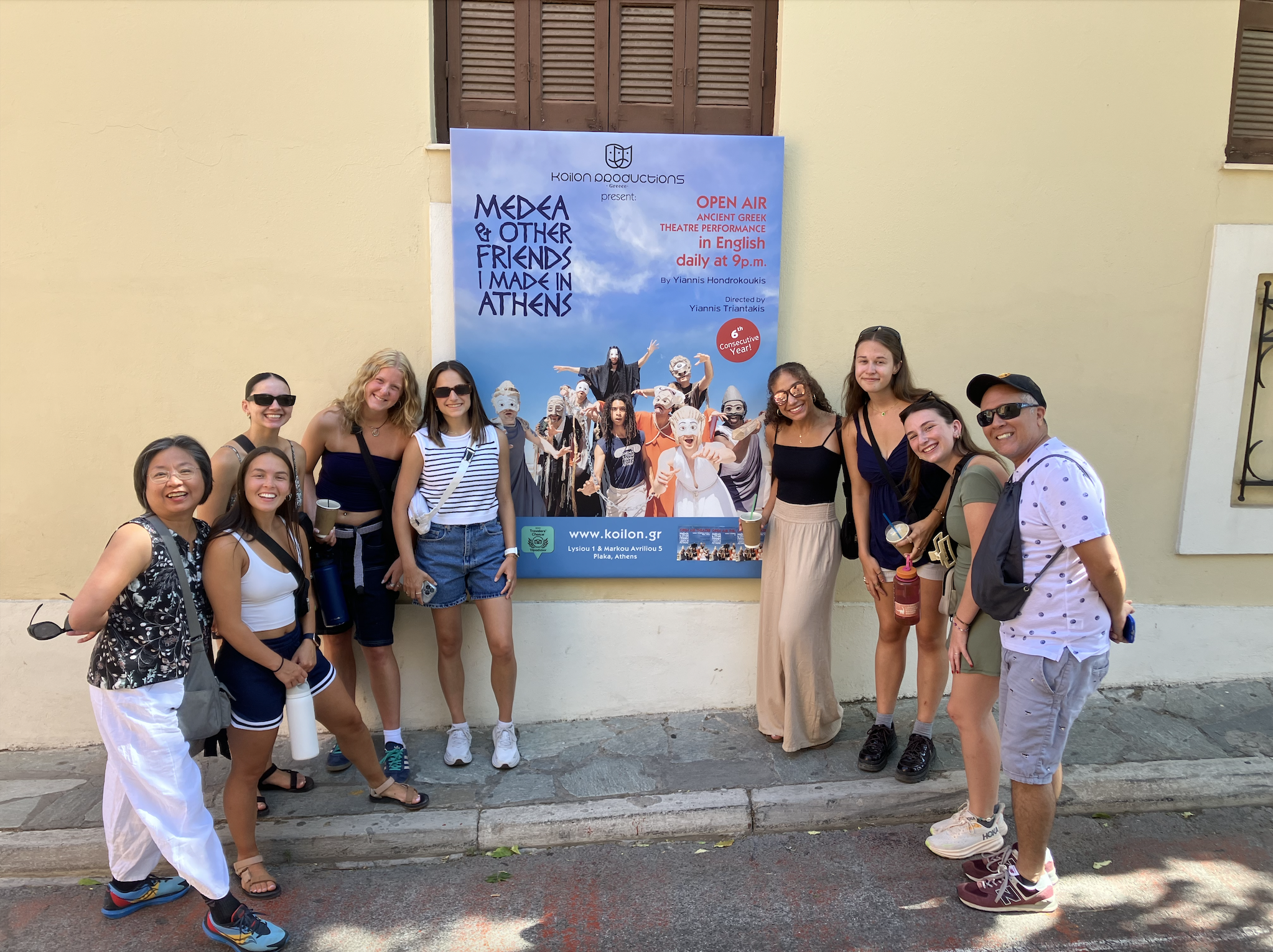 a group of students pose in front of a greek play poster outdite the stucco theater as part of a literacy scholar trip to greece from shenandoah university
