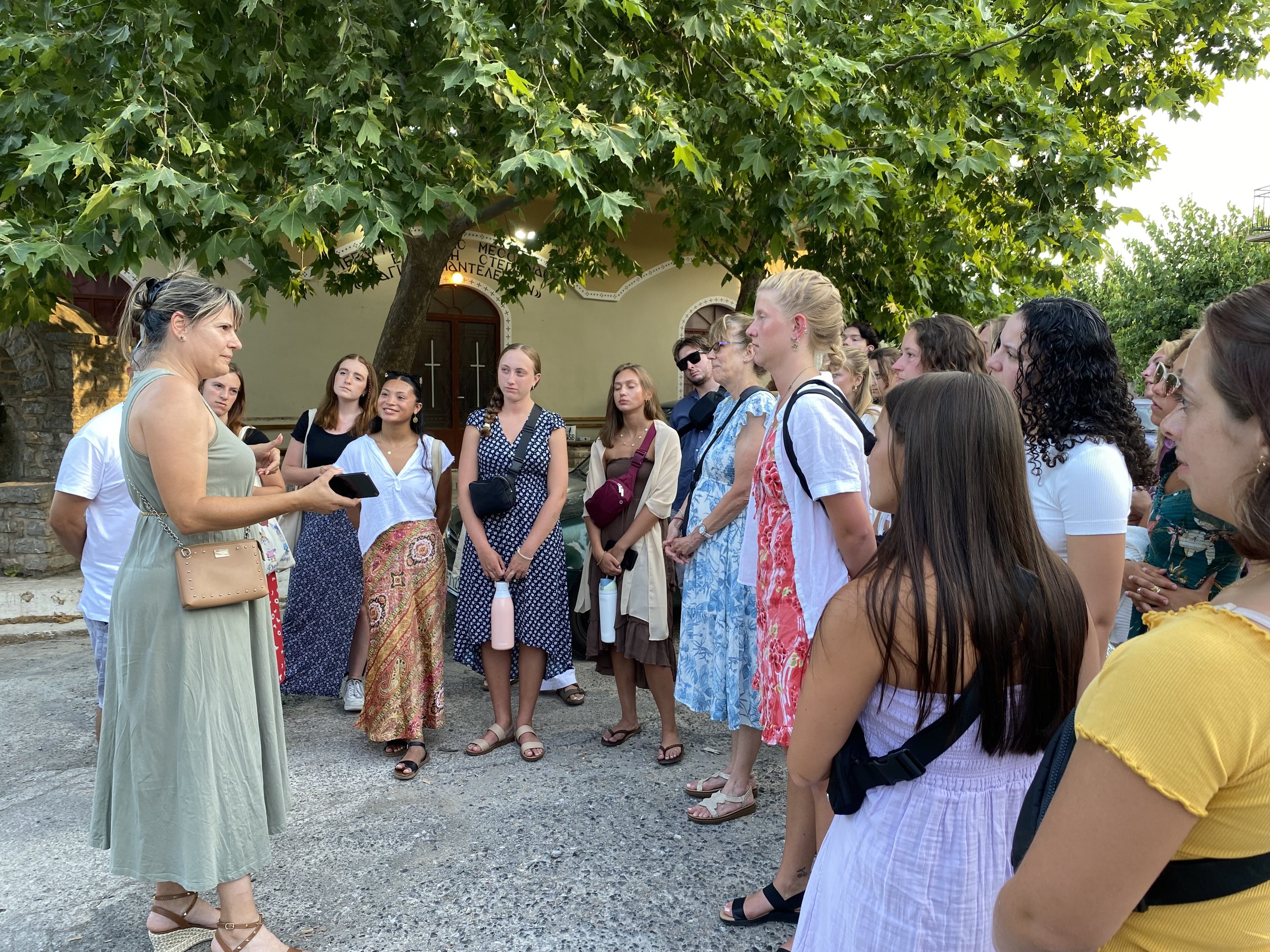 greek american scholars listen to a woman speak in a shaded patio while visiting greece to bring literacy services to rural greece. Many students are wearing sundresses and sandals. There is an oak tree shading them.