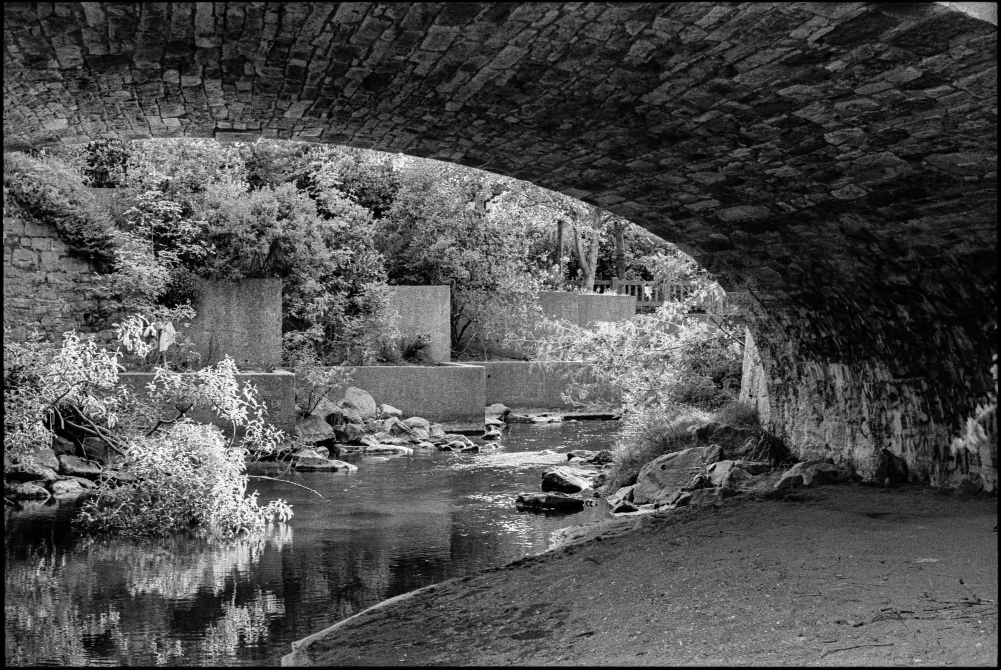 A black and white photo of a small river or stream passing underneath a stone bridge, with trees and shrubs along the banks.