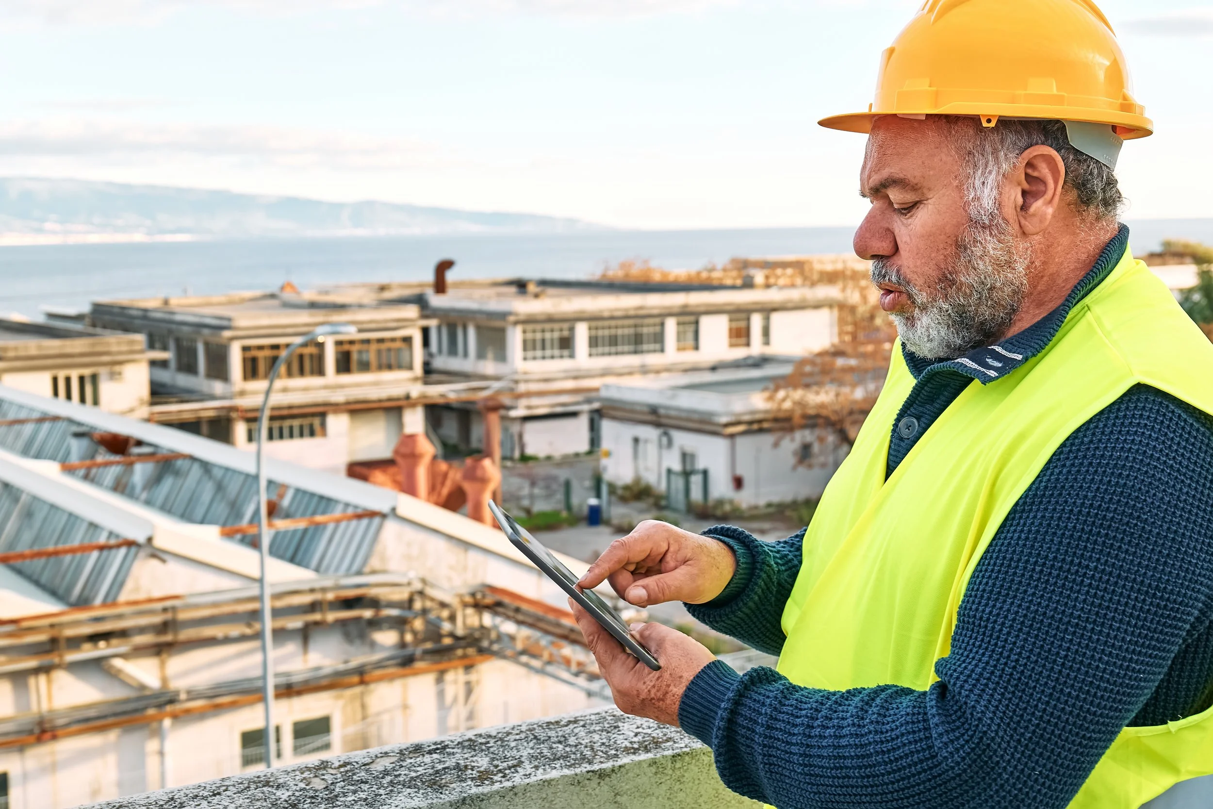 Construction worker in yellow vest and hard hat using a tablet, with industrial buildings in the background against a coastal backdrop.
