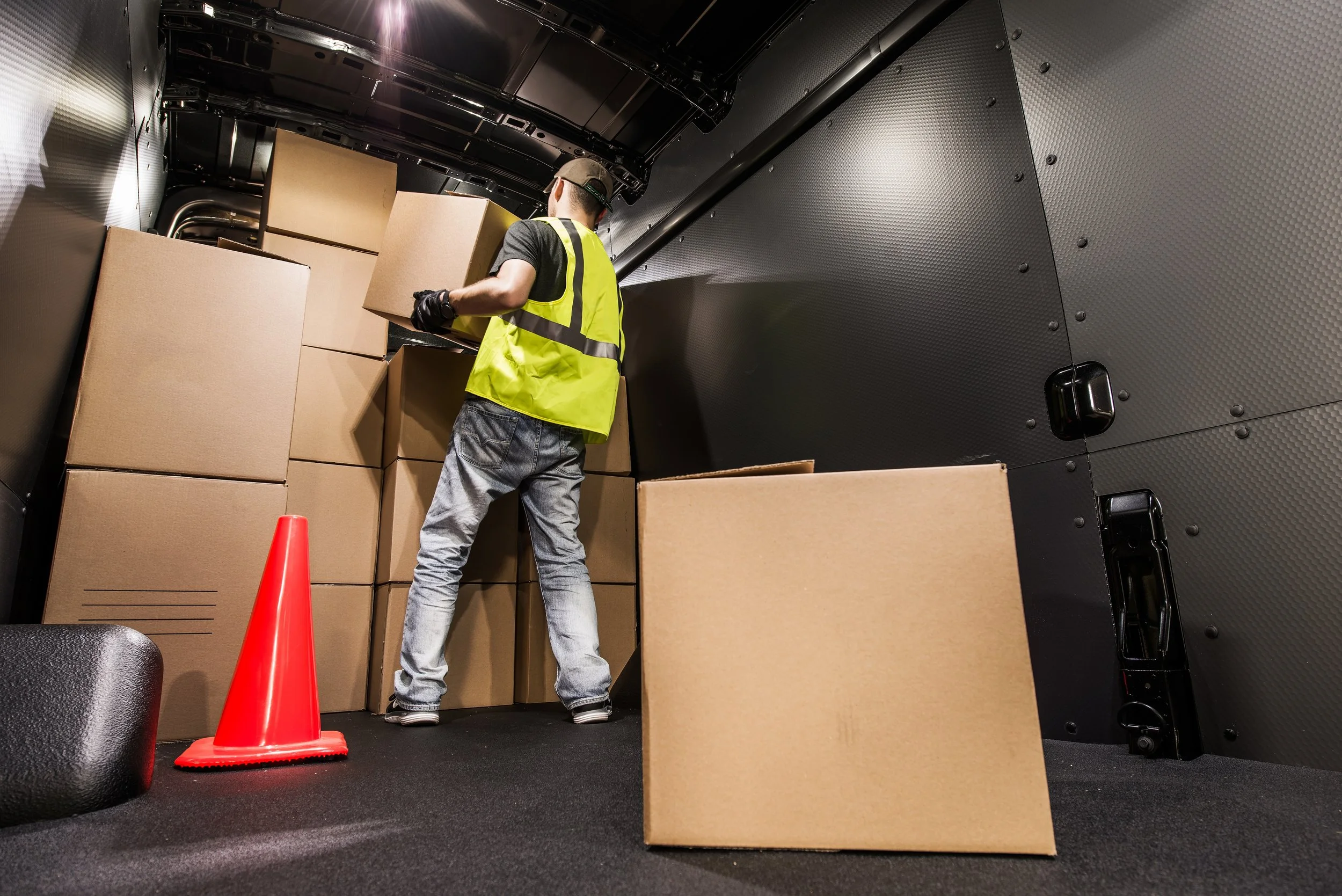 Worker in a high-visibility vest organizing cardboard boxes inside a cargo van, with an orange traffic cone on the floor.