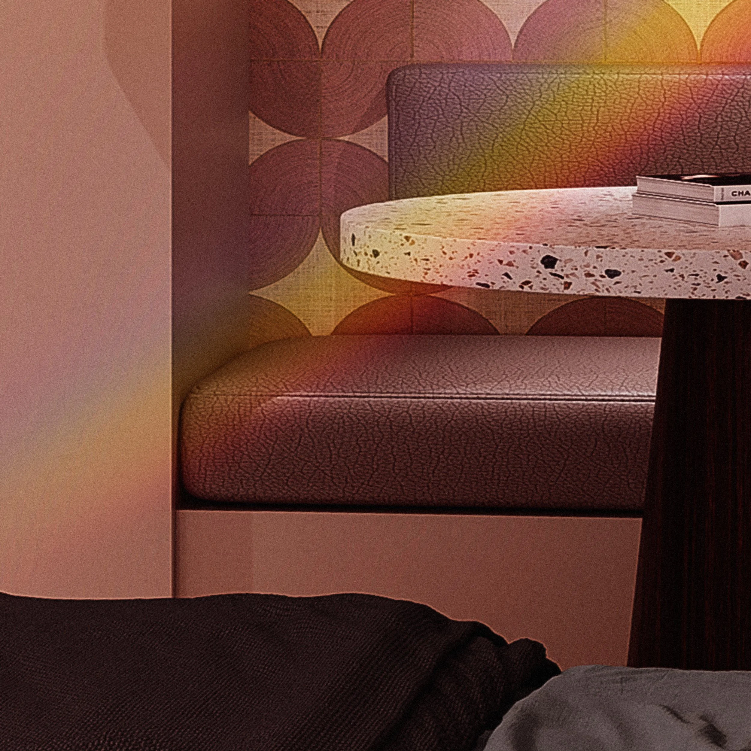 A cozy interior scene featuring a textured beige upholstered bench and a terrazzo-top round table with books on it, set against a wallpaper with a geometric pattern of overlapping circles in warm colors.