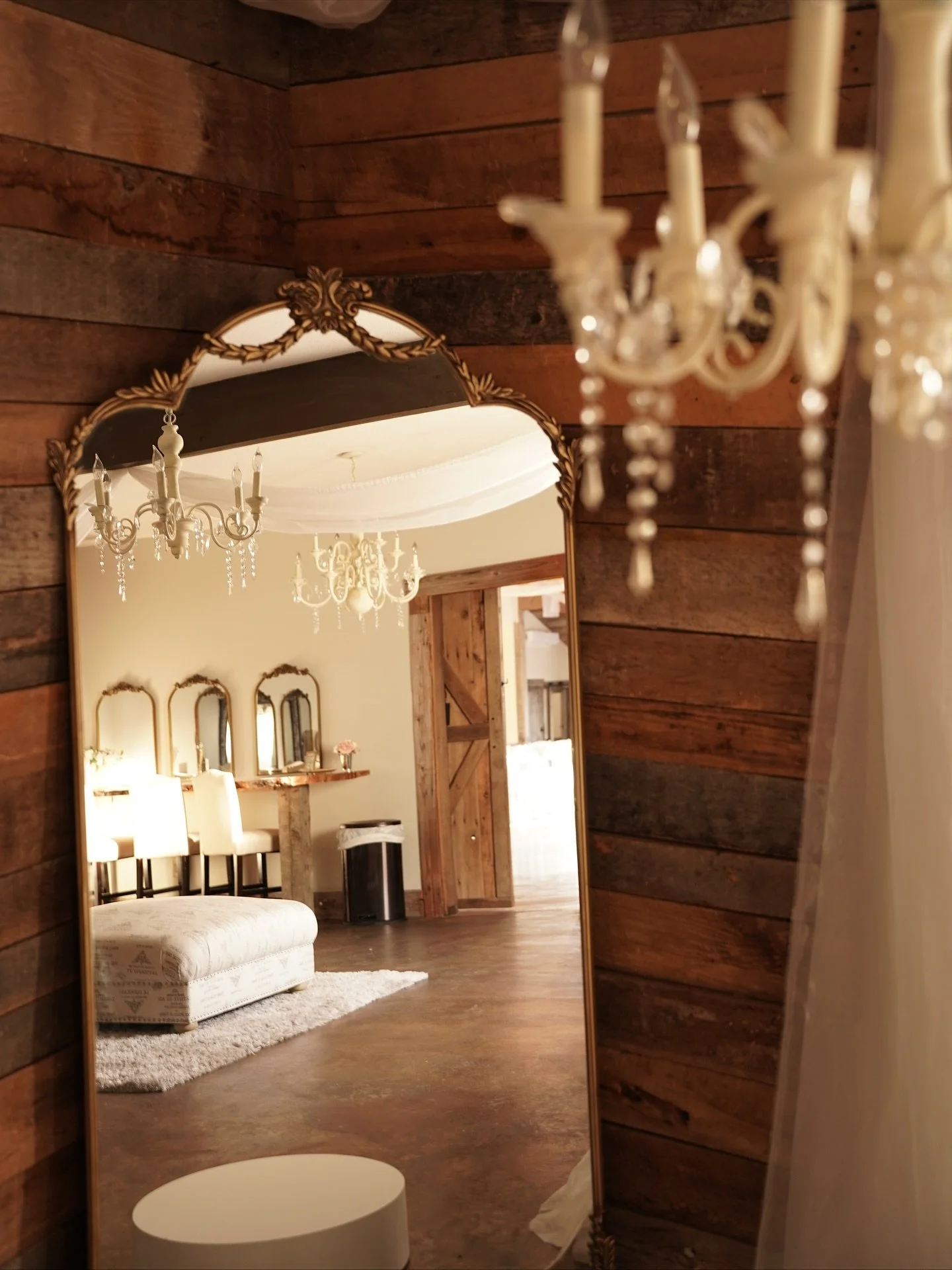 A large ornate mirror with a gold frame reflects a room with a chandelier, a cream-colored cushioned bench, a wooden wall, and a door leading outside. The room has a rustic, cozy decor with wooden accents and soft lighting.