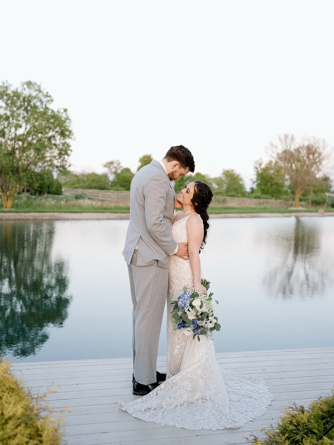 A bride and groom standing close together by a lake, sharing a moment on their wedding day. The bride holds a bouquet, and they are smiling at each other.
