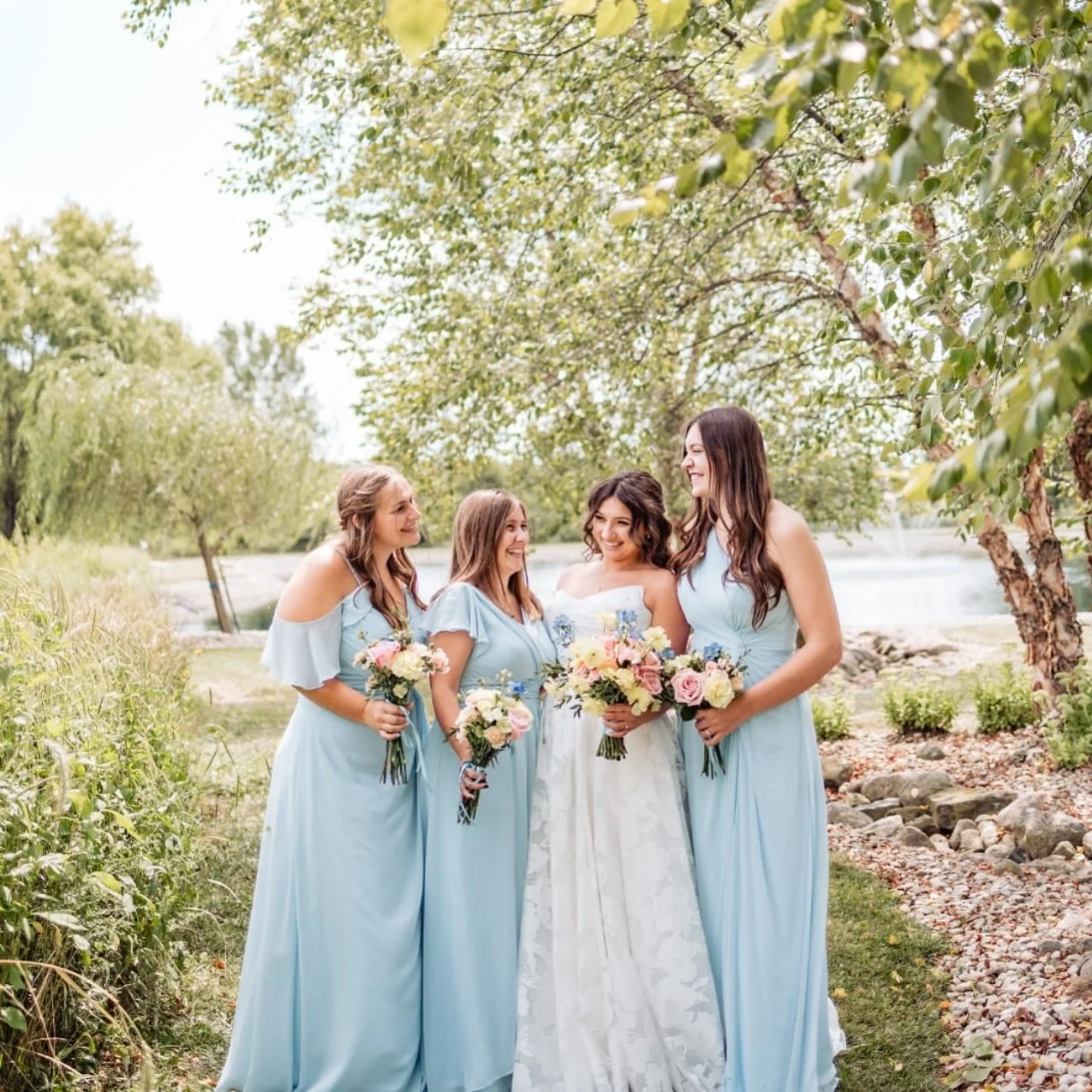 A bride and four bridesmaids standing outdoors by a river, smiling and holding bouquets of flowers, with trees and greenery in the background.