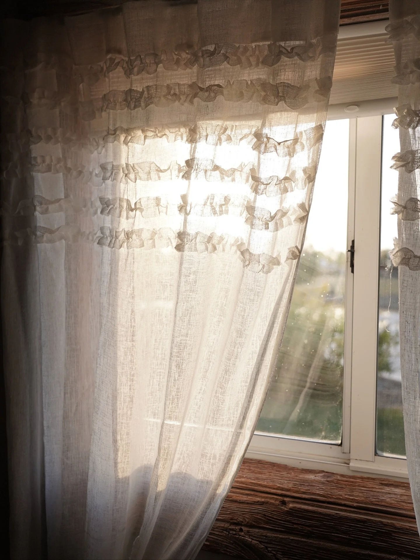 Sunlight shines through sheer, ruffled curtains on a window, revealing a glimpse of the outdoor scenery.