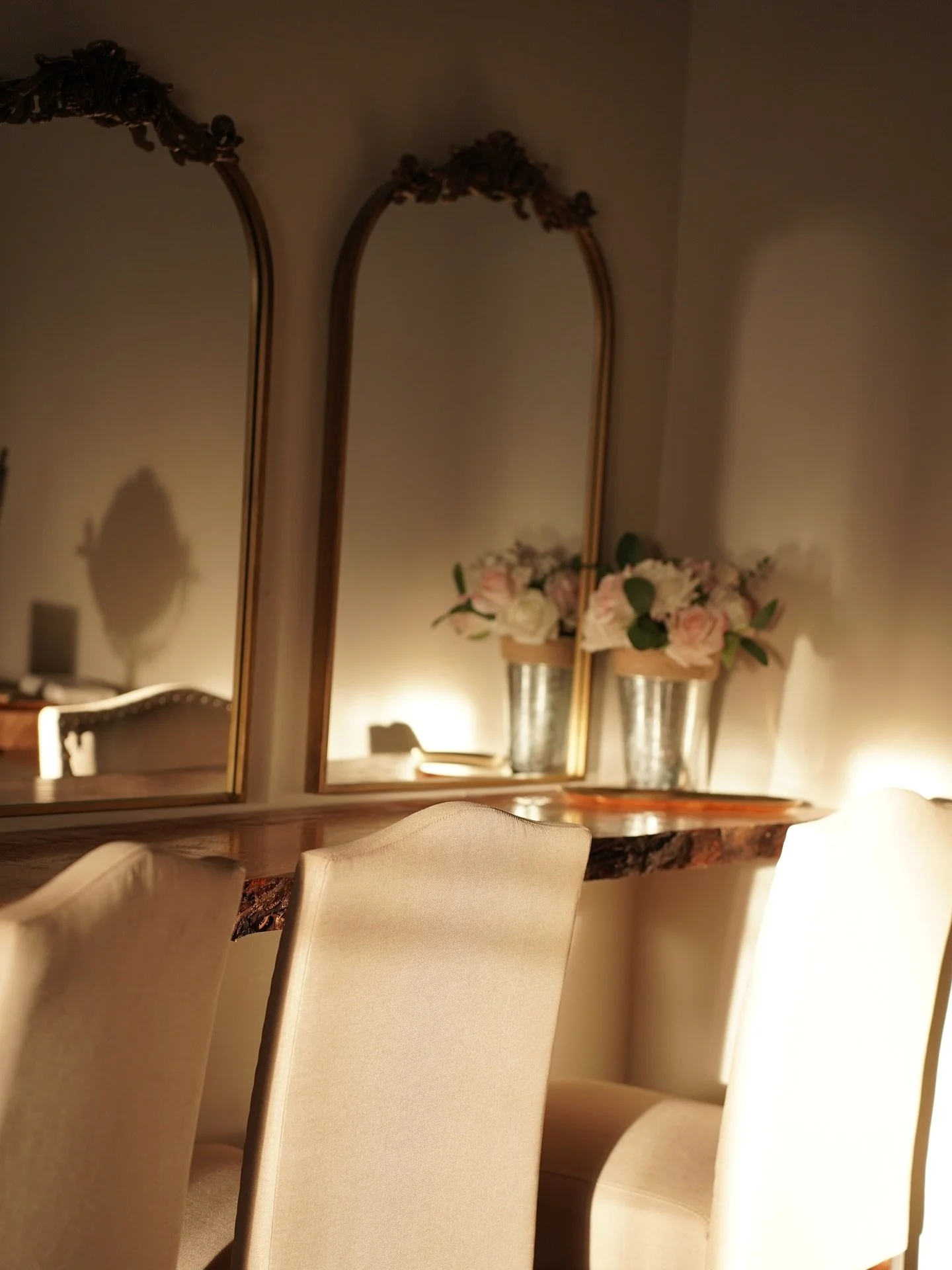 A close-up view of a dining area with beige upholstered chairs, a wooden table, and a large gold-framed mirror. Two silver vases with pink and white flowers are on the table, reflected in the mirror, with soft warm lighting creating a cozy atmosphere