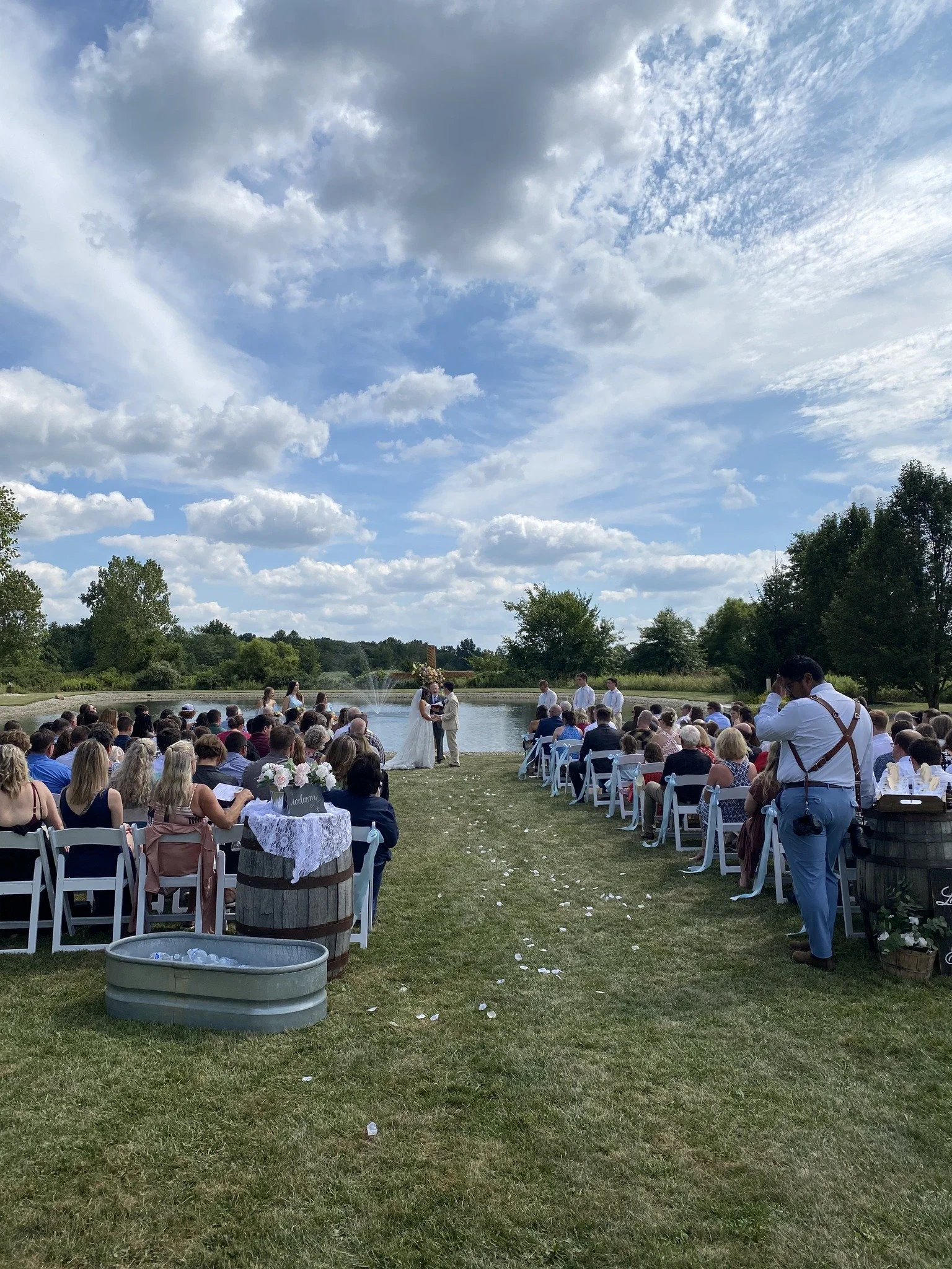 Outdoor wedding ceremony with guests seated on white chairs on a grassy area by a pond, with a bride and groom kissing at the altar under a partly cloudy sky.